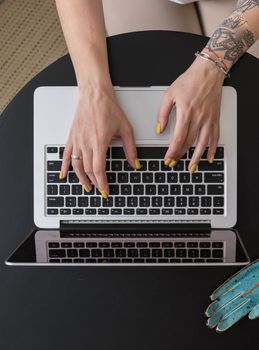 Top view of tattooed hands typing on a laptop with yellow polished nails.