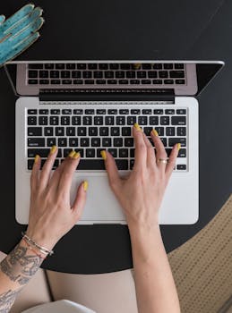 Close-up of hands with yellow manicured nails typing on a laptop keyboard from an overhead angle.