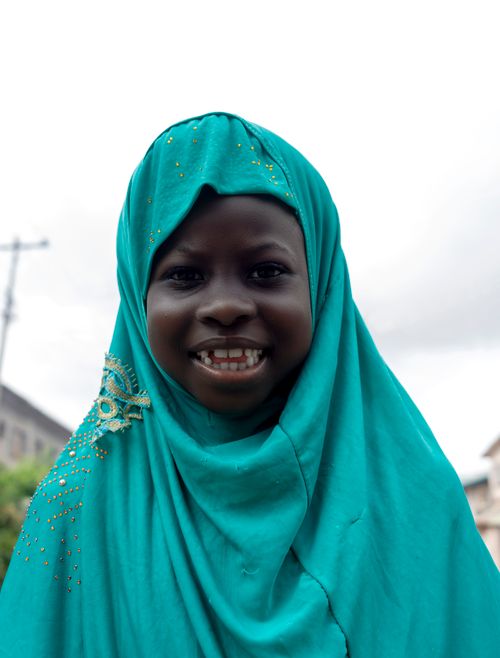 Free A cheerful young girl wearing a decorated teal hijab smiles warmly outdoors against a blurred background. Stock Photo