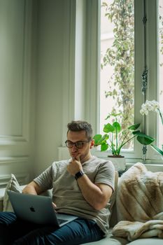 A man wearing eyeglasses using a laptop in a cozy home setting with natural light.