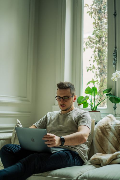 Free A man using a laptop in a bright, modern living room with a large window. Stock Photo