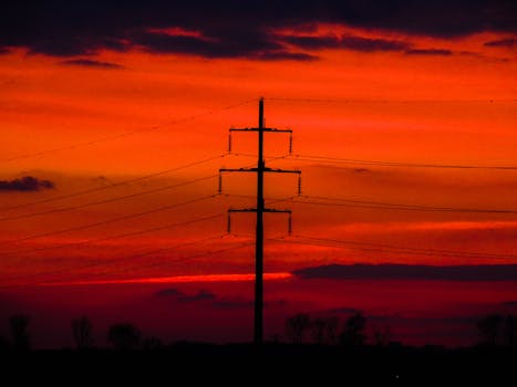 Silhouette of power lines against a vibrant red and orange sunset sky, creating a dramatic energy landscape.
