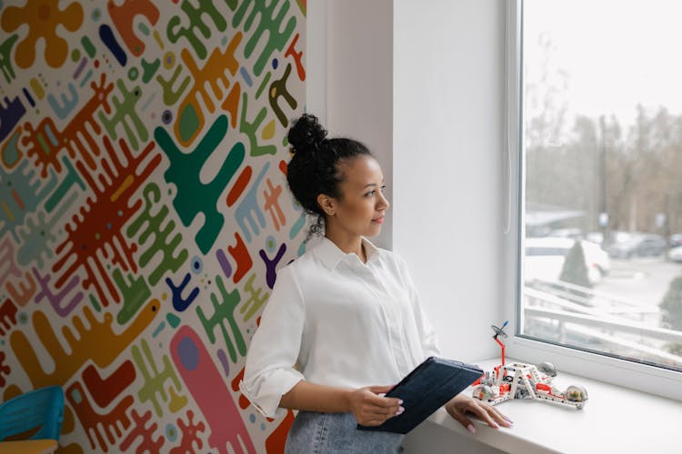 A Woman In White Long Sleeves Holding A Tablet While Looking At The Window