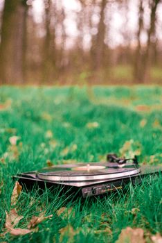A vintage turntable playing a record amidst fallen leaves and grass in an autumn forest setting.