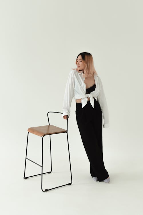 A Woman in White Long Sleeve Shirt and Black Pants Standing Beside a Chair