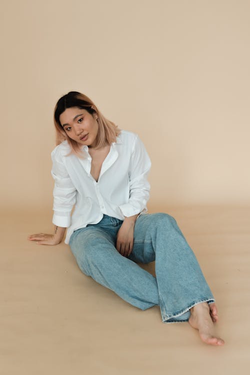 Woman in White Dress Shirt and Blue Denim Jeans Sitting on Brown Floor