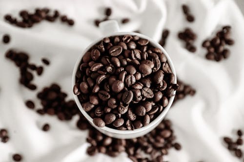 Top view of coffee beans in cup near scattered grains on crumpled white cloth