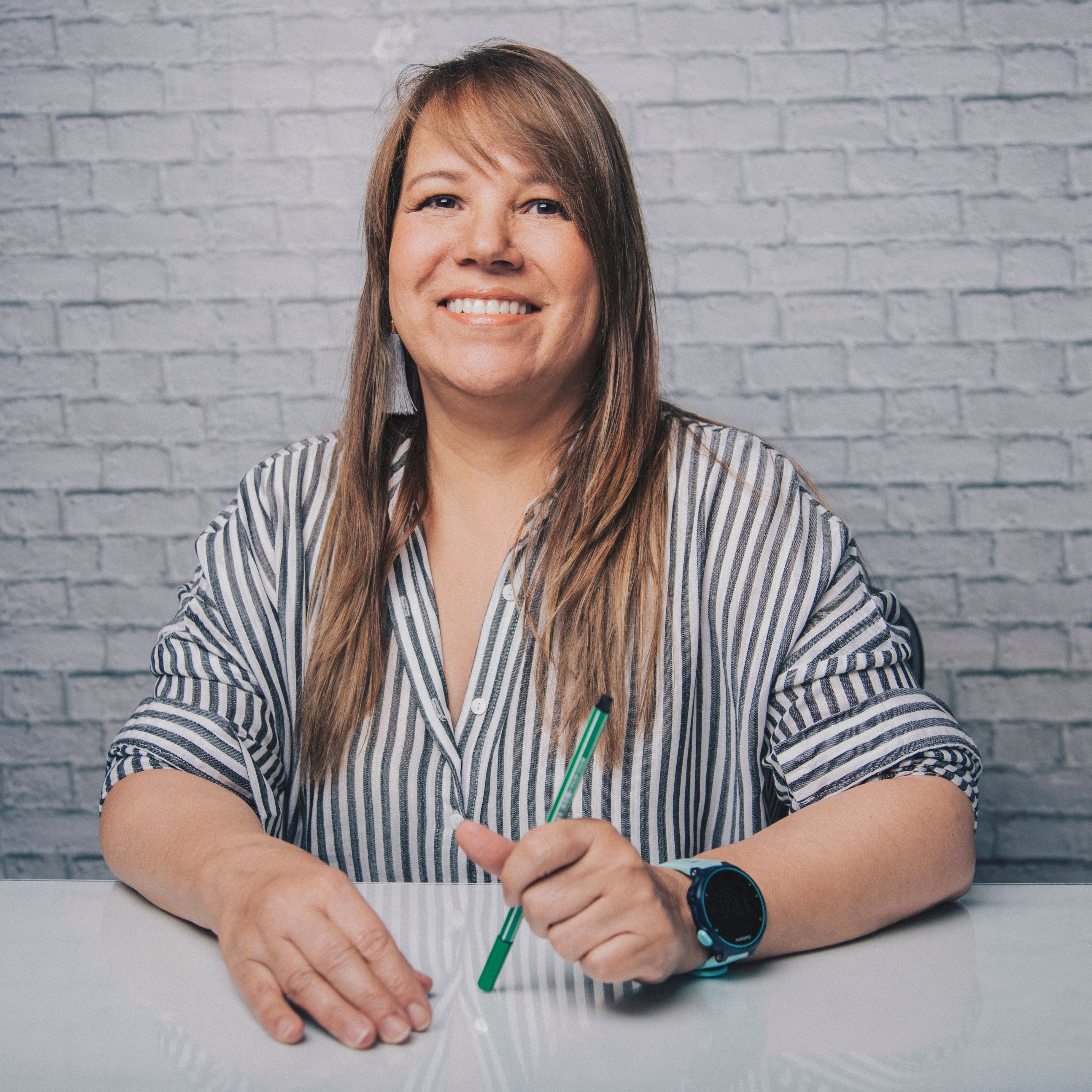 Happy woman in casual clothes with watch and earrings sitting at table and holding pen while smiling and looking away in studio with white brick wall
