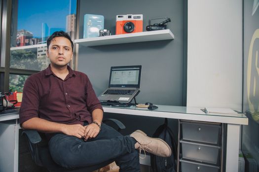 Serious male in casual clothes sitting with crossed legs near desk with laptop and shelf while looking at camera in modern workplace