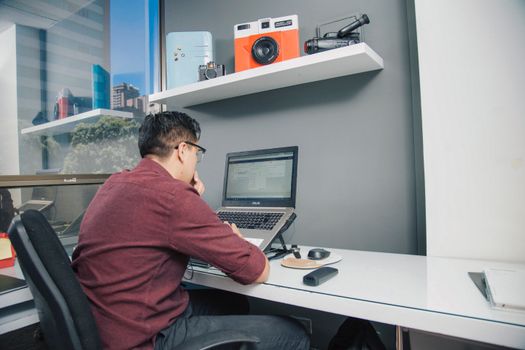 Man in maroon shirt working on laptop at a modern office desk.