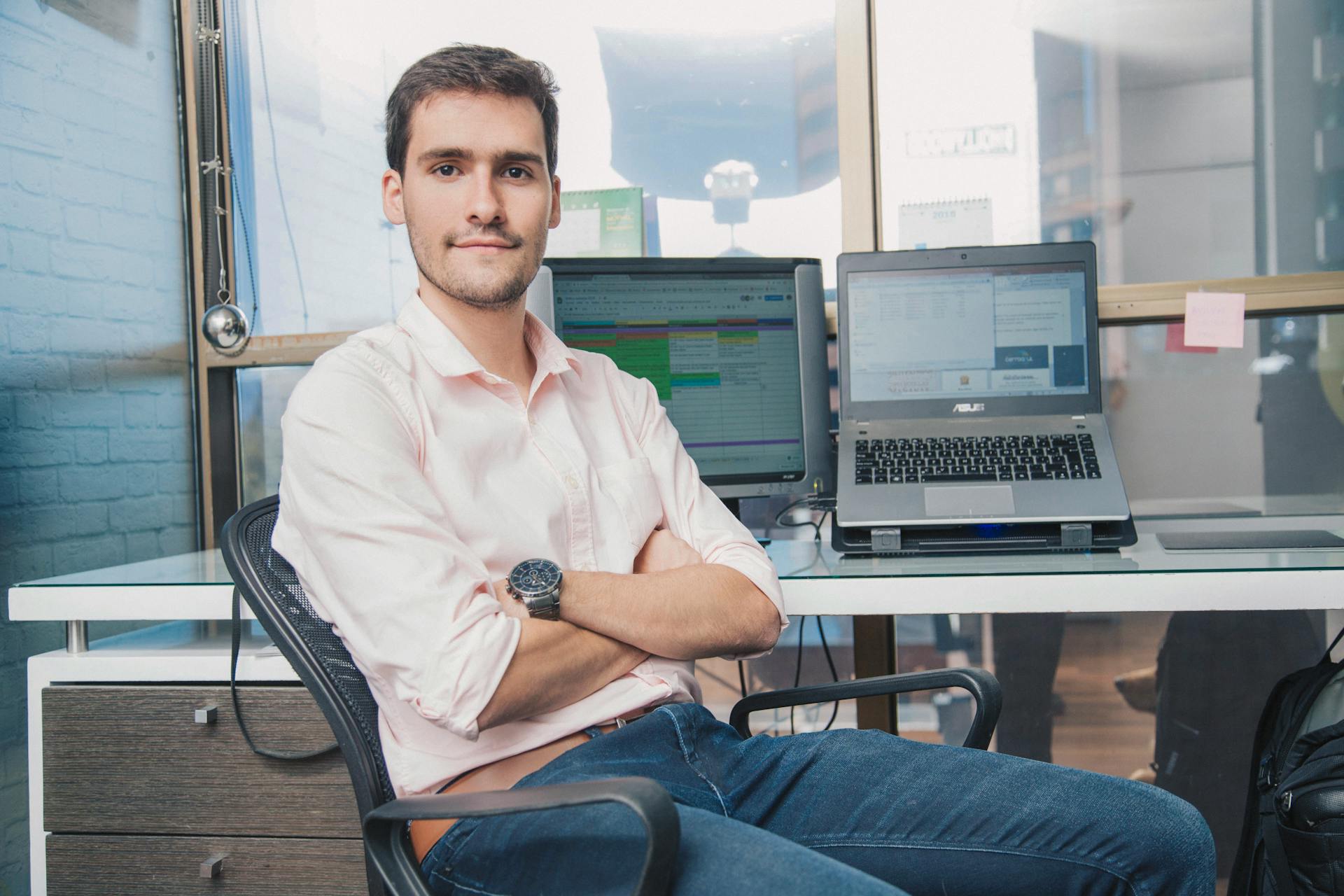 Confident young man seated in a modern office setting with laptops and screens around him.