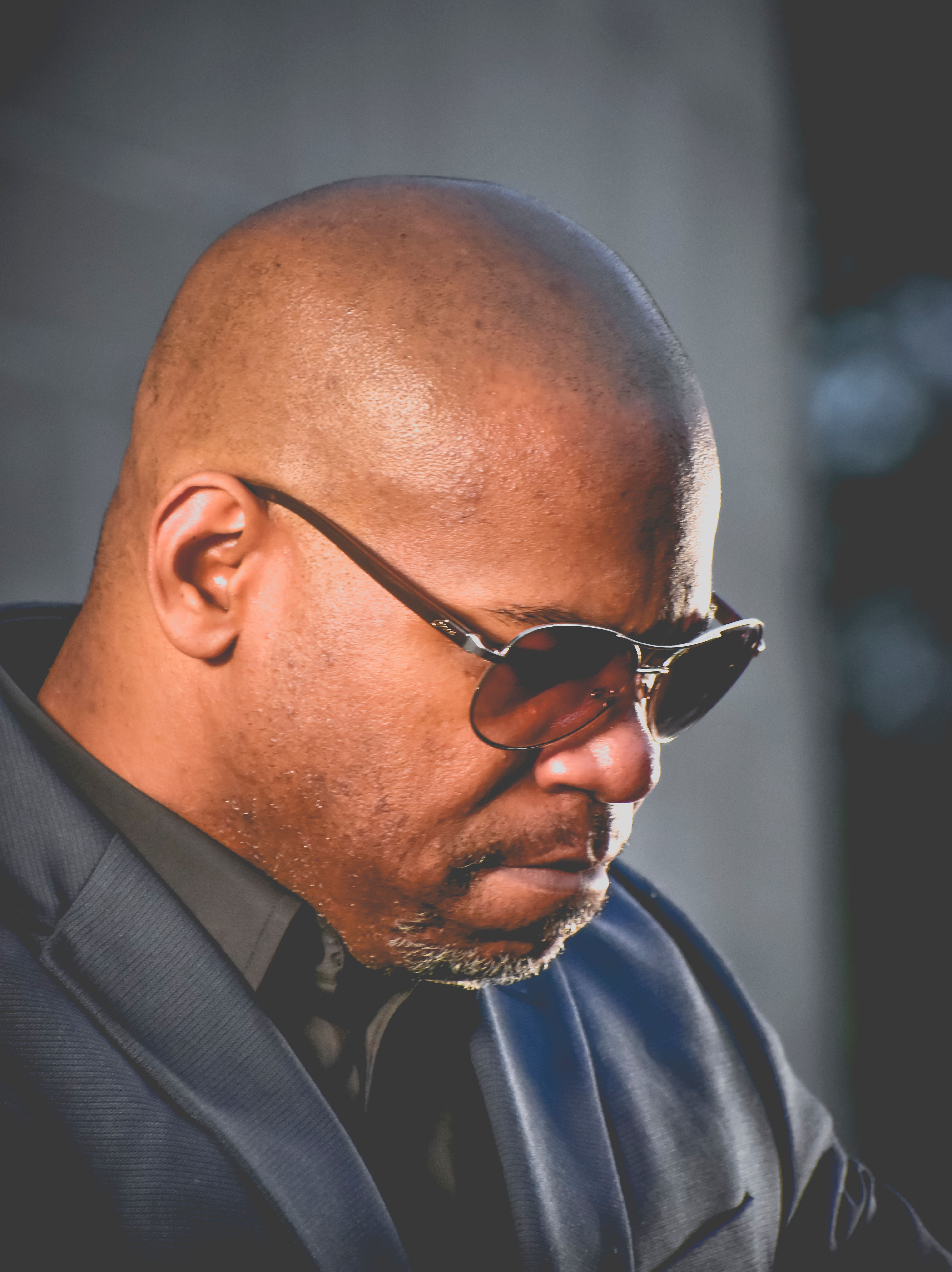 Thoughtful bald man wearing sunglasses and suit jacket looking downward.