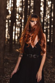 A woman with long red hair and a black dress poses in an enchanting forest setting.