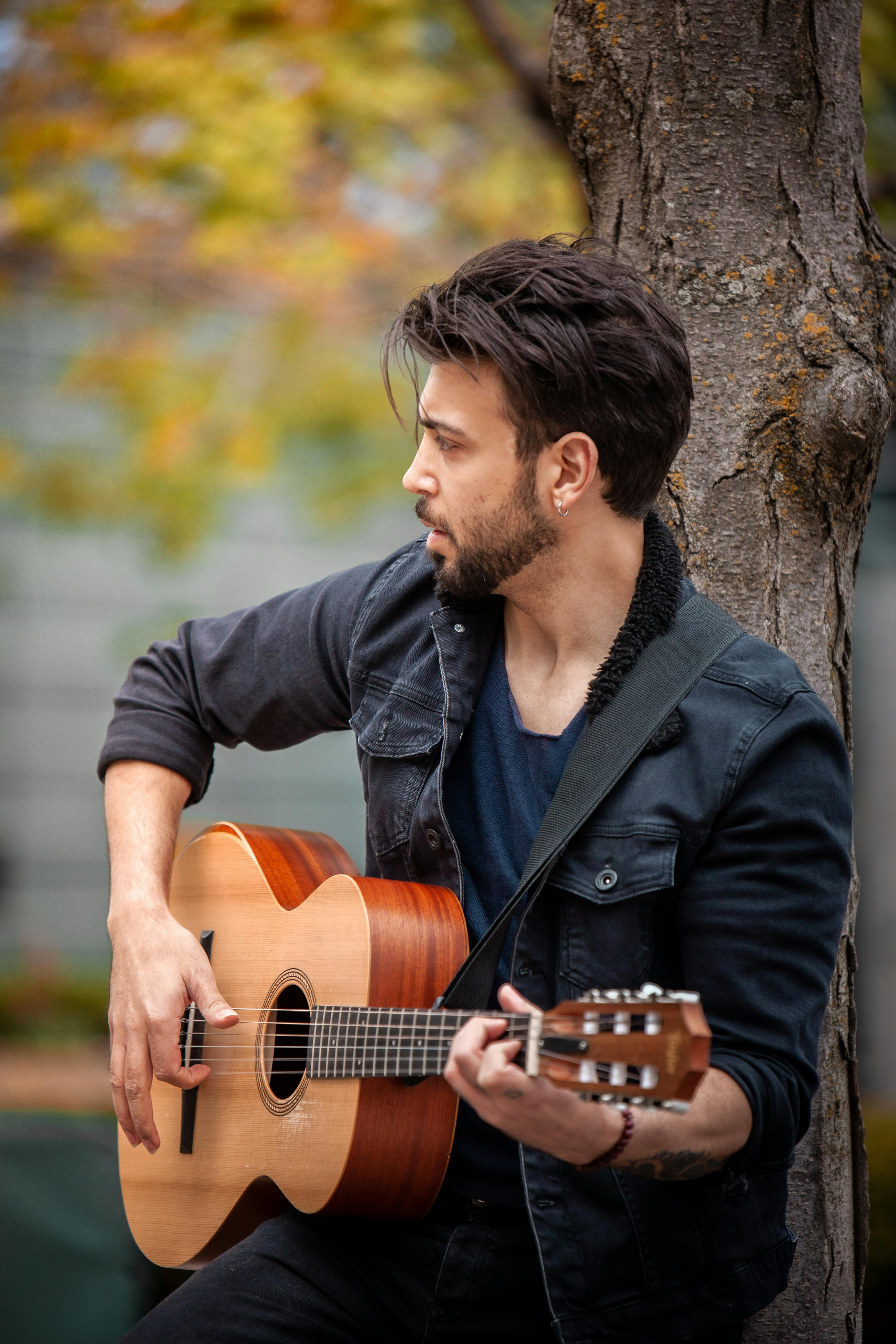 A bearded man enjoys playing guitar while leaning against a tree outdoors.