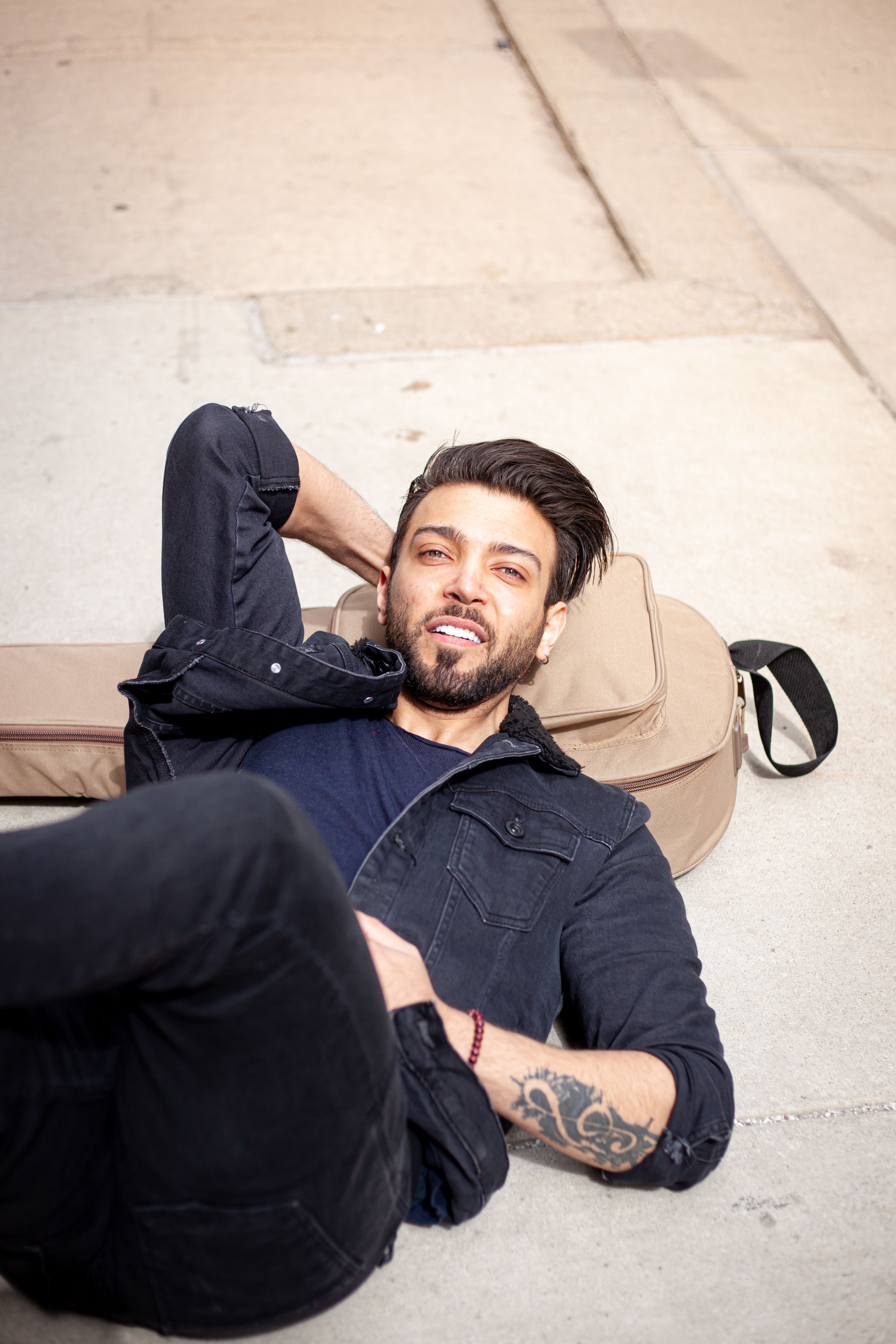 Tattooed man lying on ground, smiling beside guitar bag. Urban lifestyle scene.