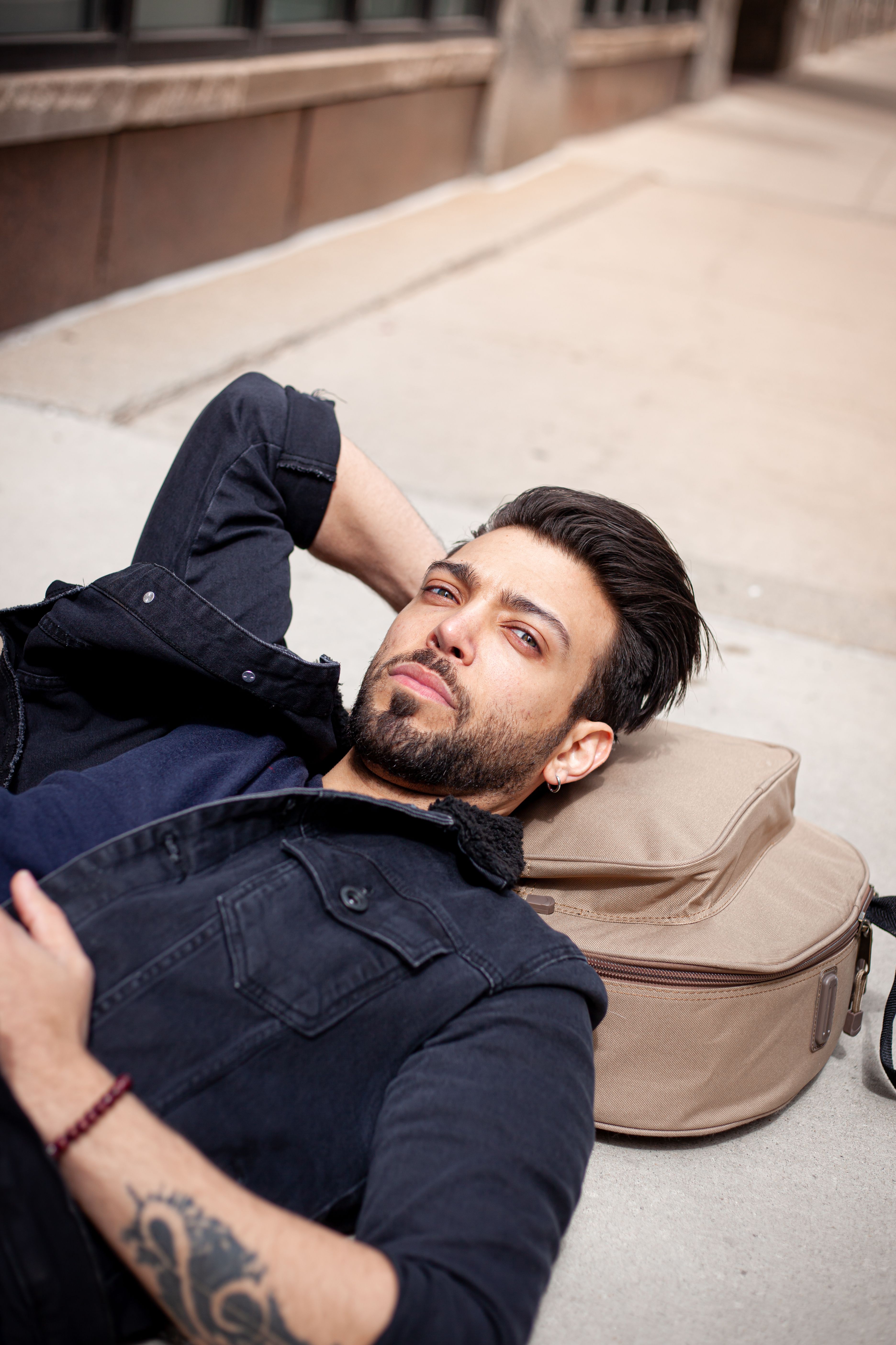 A stylish man with a beard relaxes on a city sidewalk, exuding confidence.