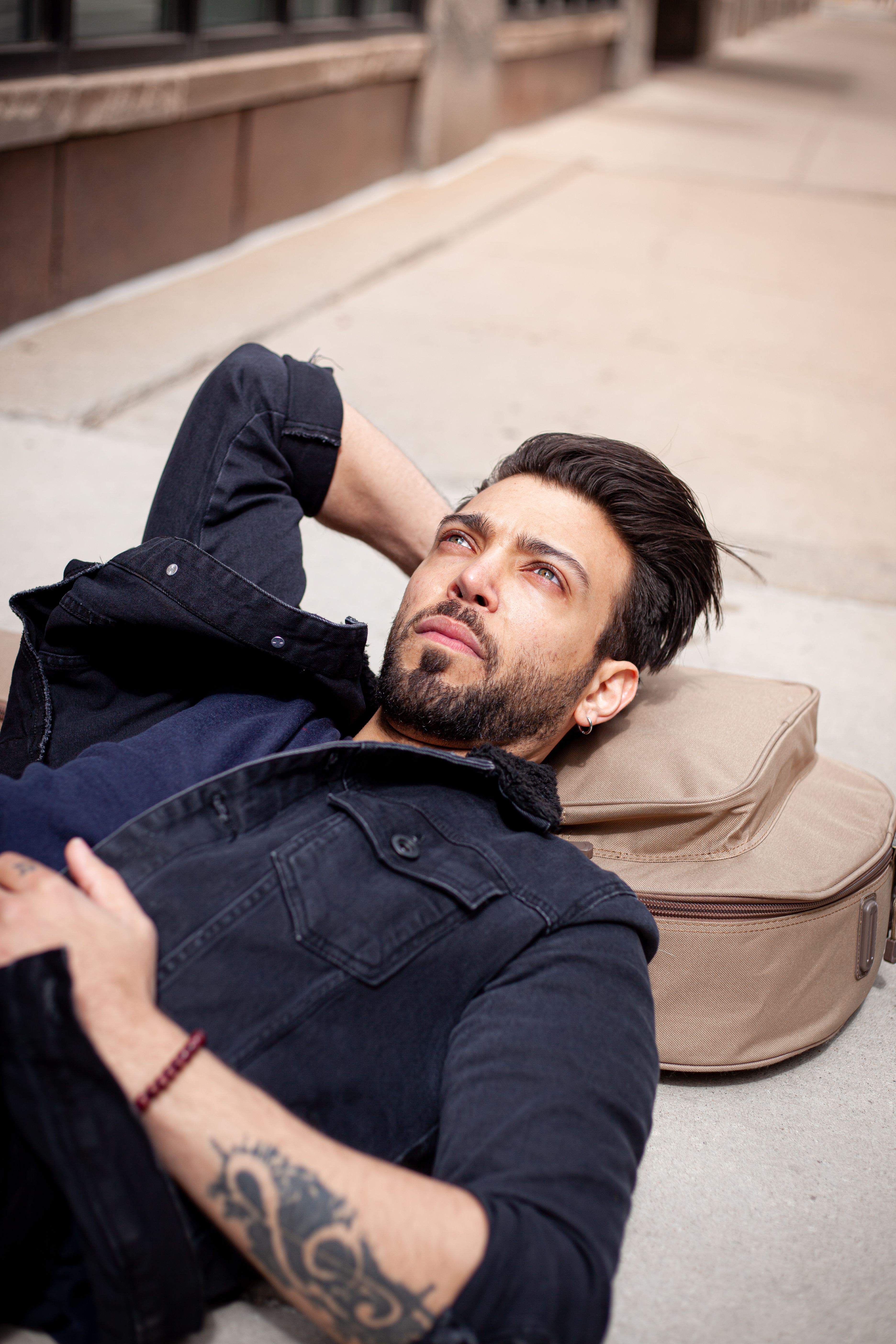 A bearded man with tattoos lies on the sidewalk, gazing thoughtfully.