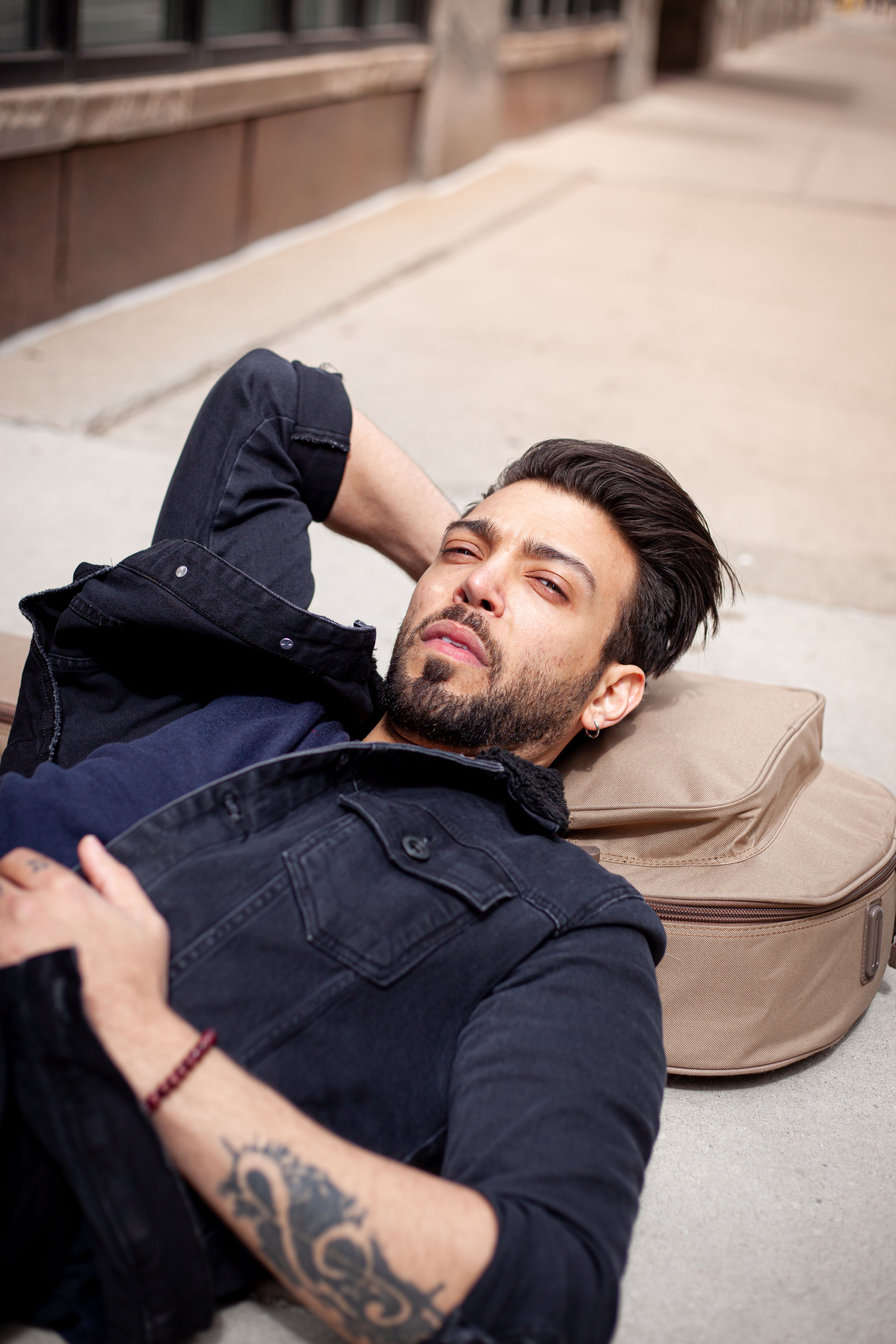 A fashionable man in a black jacket relaxes outdoors on the sidewalk.