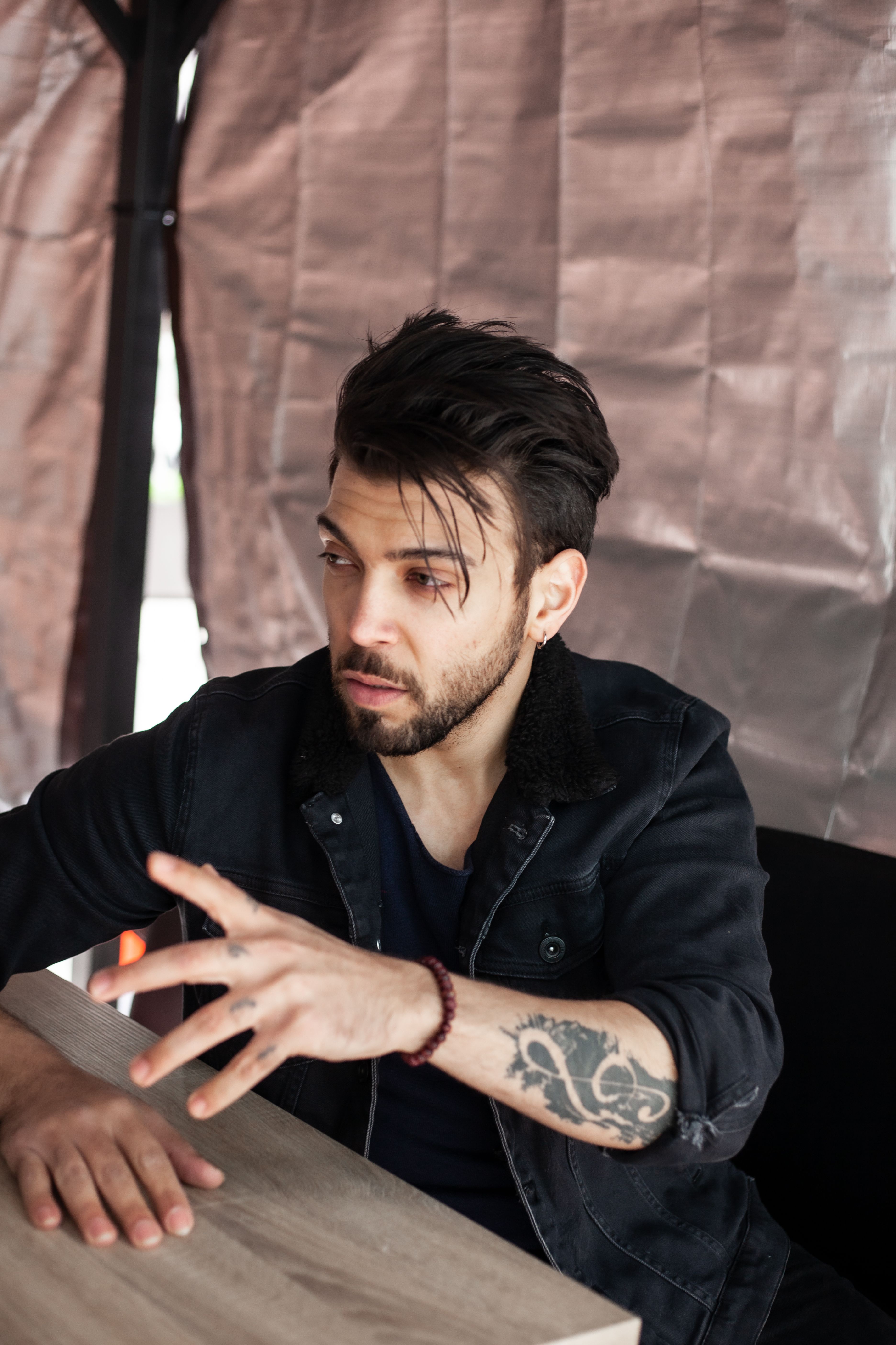 Bearded man with a tattoo in a casual indoor setting, gesturing with his hands.