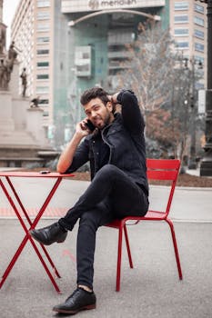 Stylish man in denim jacket sitting and talking on a smartphone by a red table outdoors.