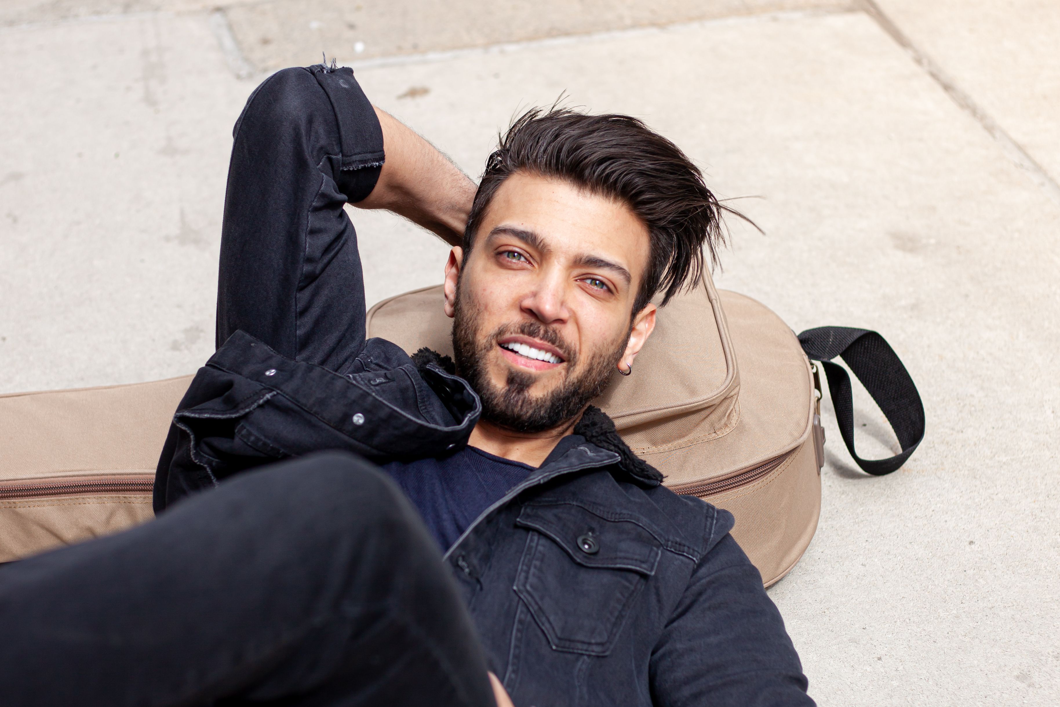 A man lying on the ground smiling, dressed in casual black denim with a guitar case outdoors.