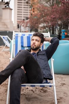 A bearded man in a denim jacket relaxes on a beach chair in an urban setting.