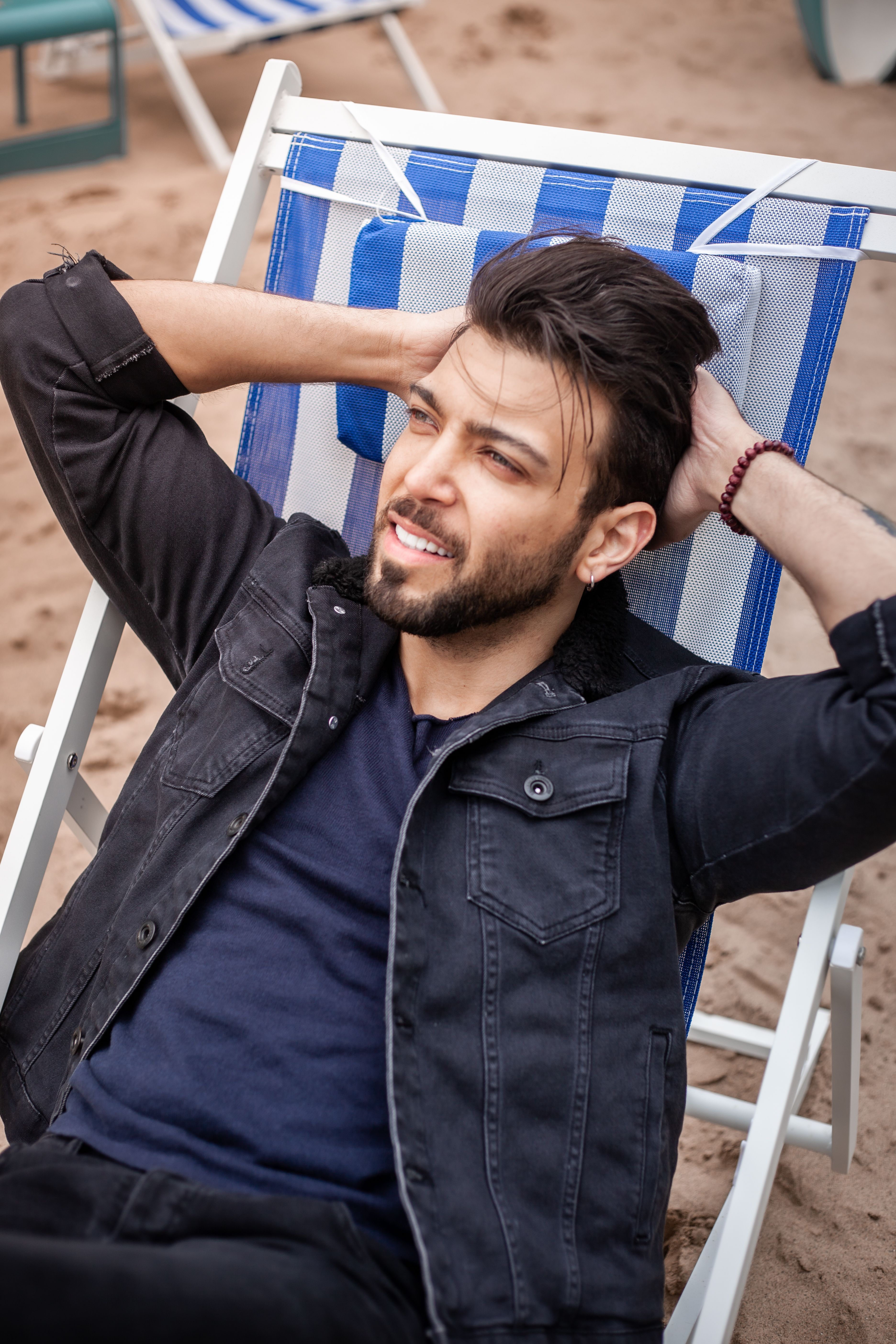 Man in a denim jacket relaxing on a beach chair with a smile, enjoying a sunny day outdoors.