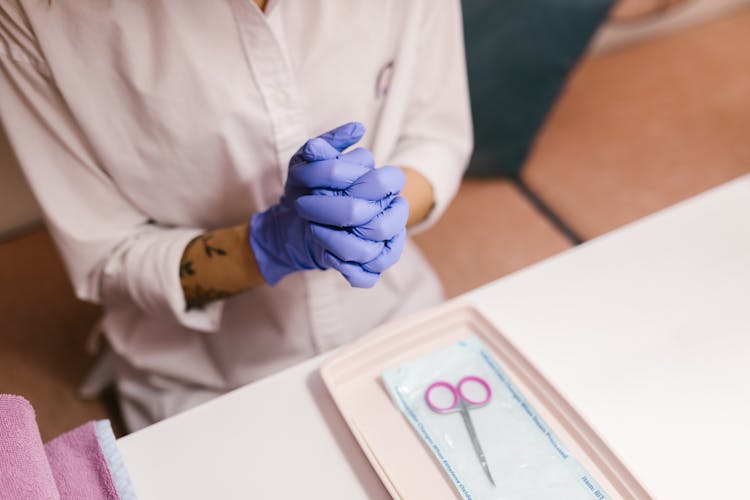 Person In Blue Gloves With Hands Clasped Sitting Behind A White Table With Scissors