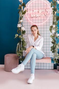 Stylish woman sitting on a swing with neon sign, exuding modern elegance and charm.