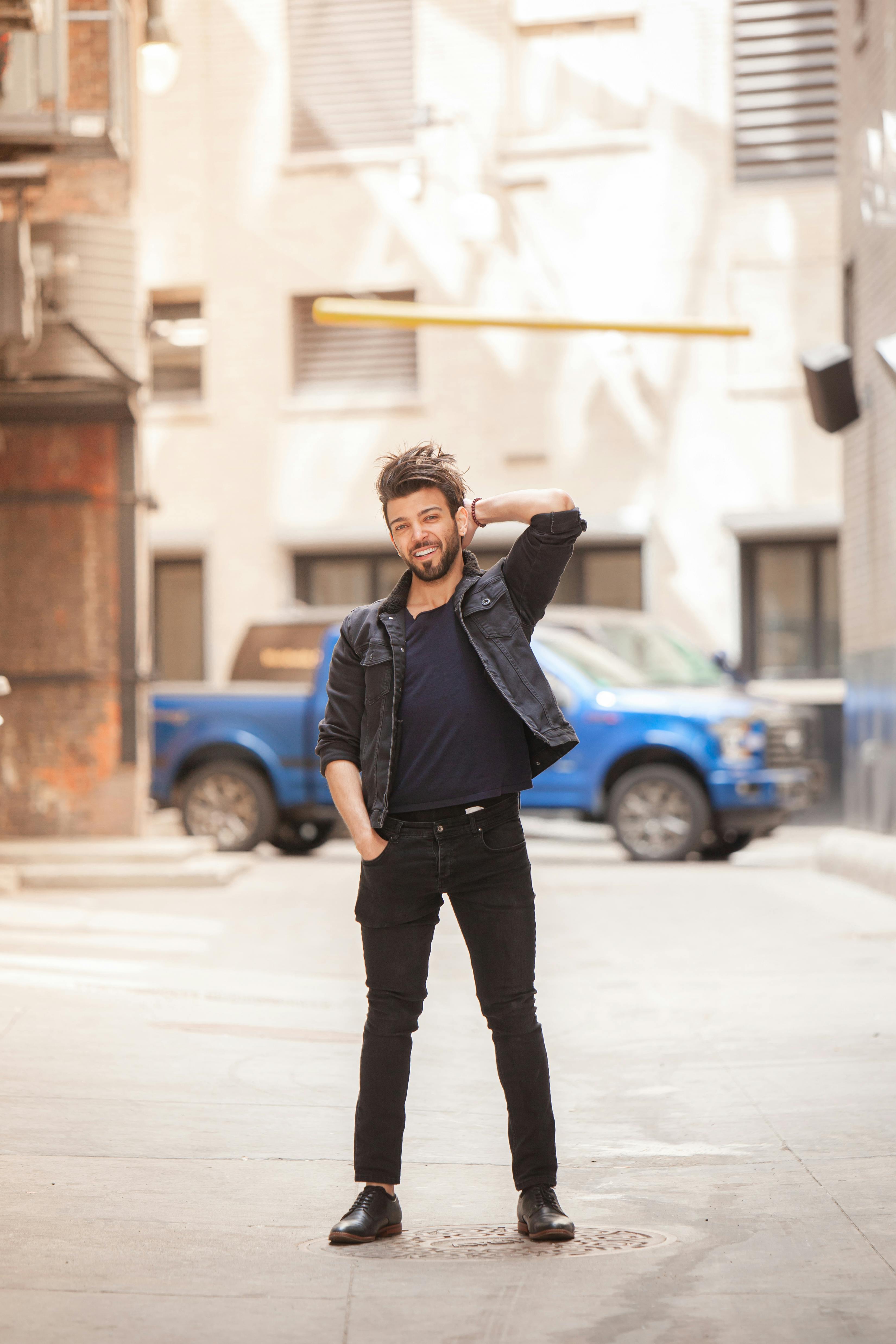 A bearded man in black attire poses confidently in a bright urban alley with blue truck.