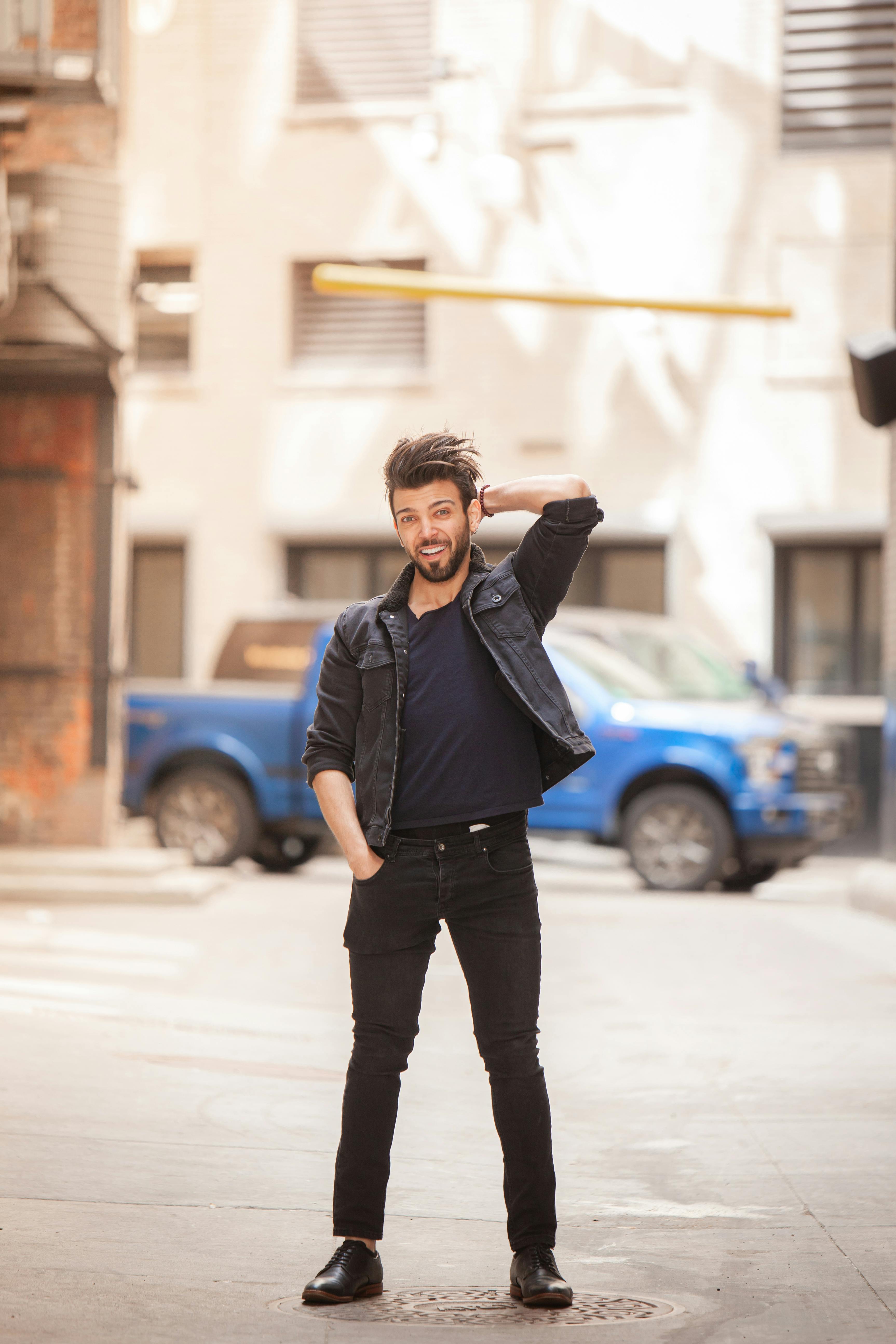 A bearded man in a denim jacket smiling while posing on an urban street.