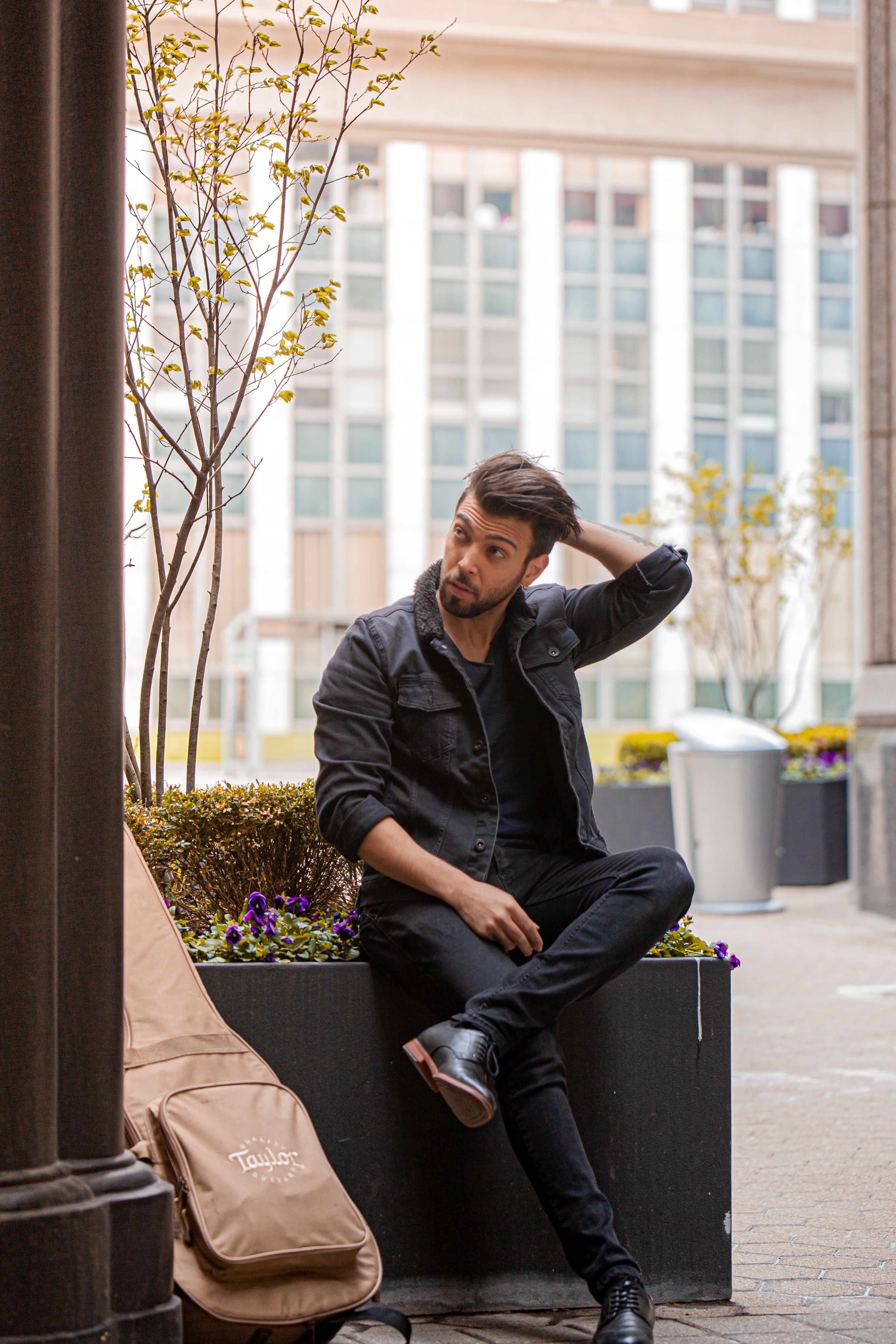 Man in casual fashion sitting in a city area with plants and guitar case nearby.