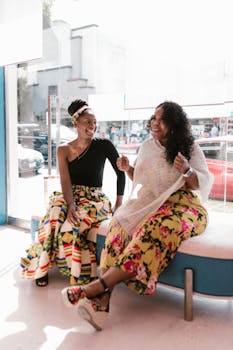 Two stylish black women sharing a joyful moment at a trendy salon, with sunlight streaming through large windows.