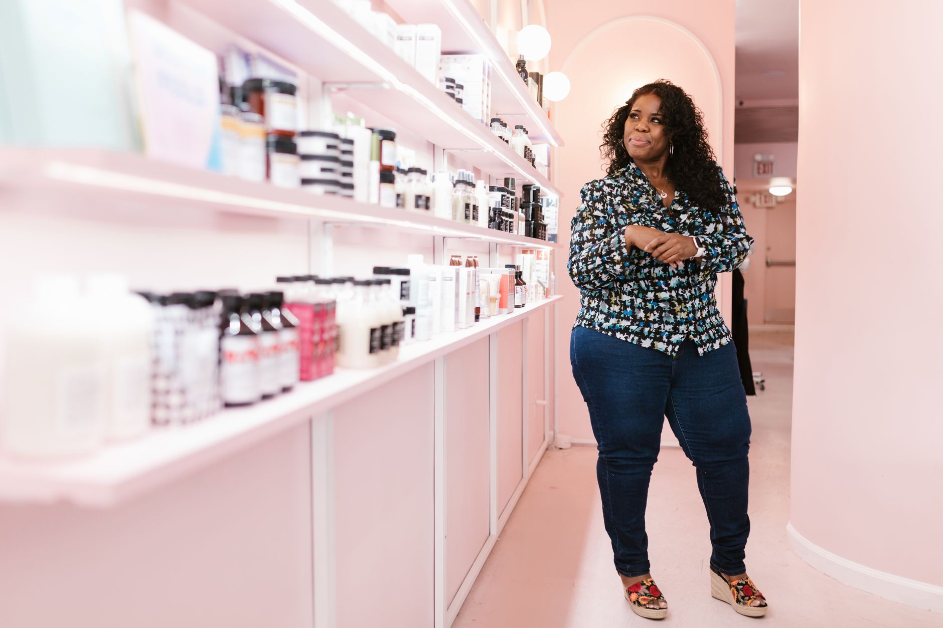 Woman standing in a modern beauty shop, smiling confidently beside shelves of cosmetics.