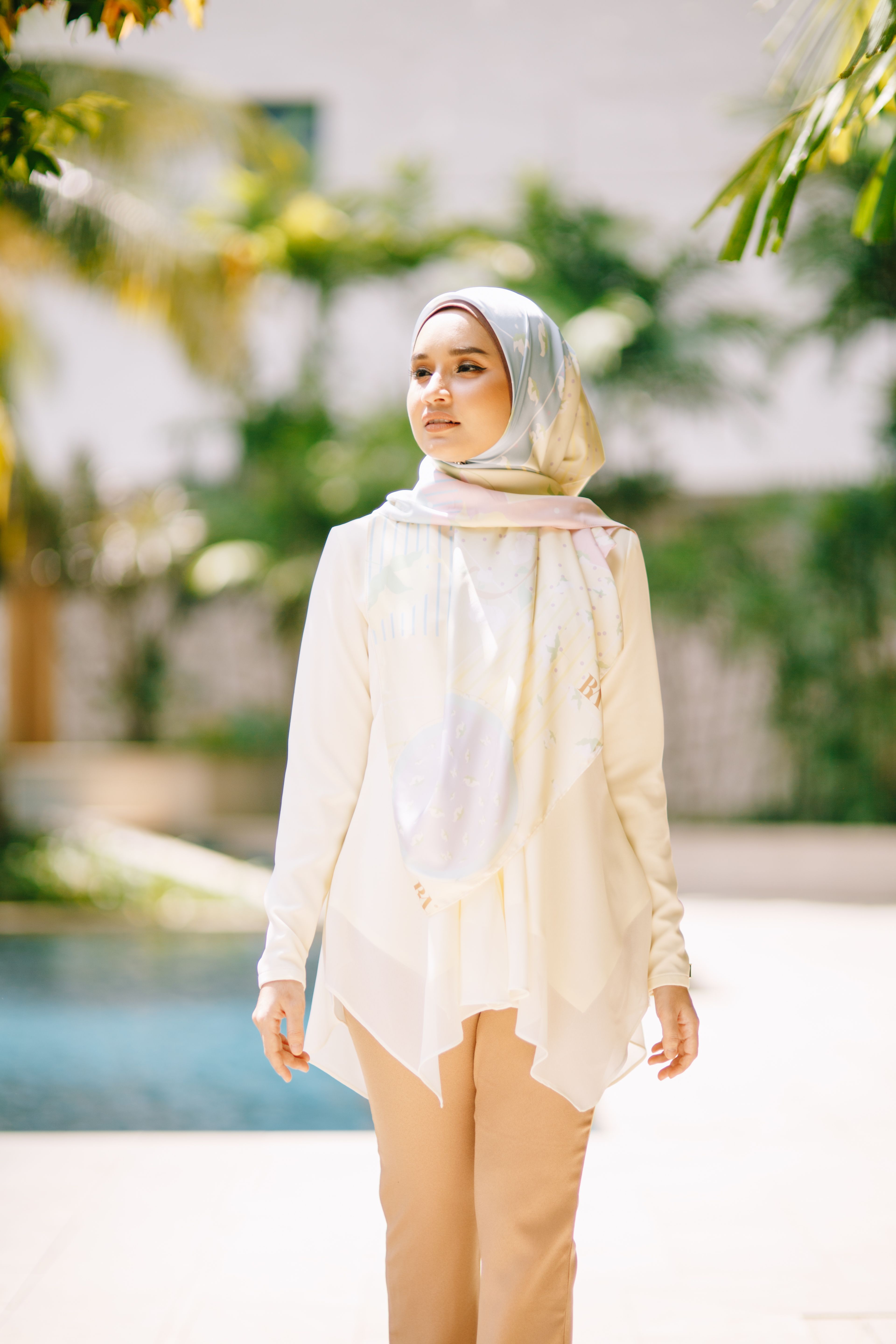 Portrait of a young Asian woman in a white blouse and hijab, walking outdoors on a sunny day.