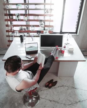 Man in casual setting reading book, feet on desk, showcasing relaxed home office vibe.