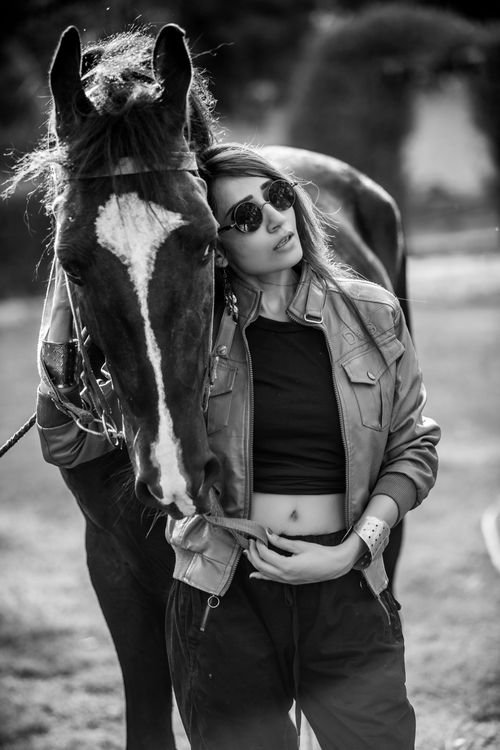Free Black and white shot of young attractive female standing near horse in deserted park and wearing eyeglasses and leather jacket Stock Photo