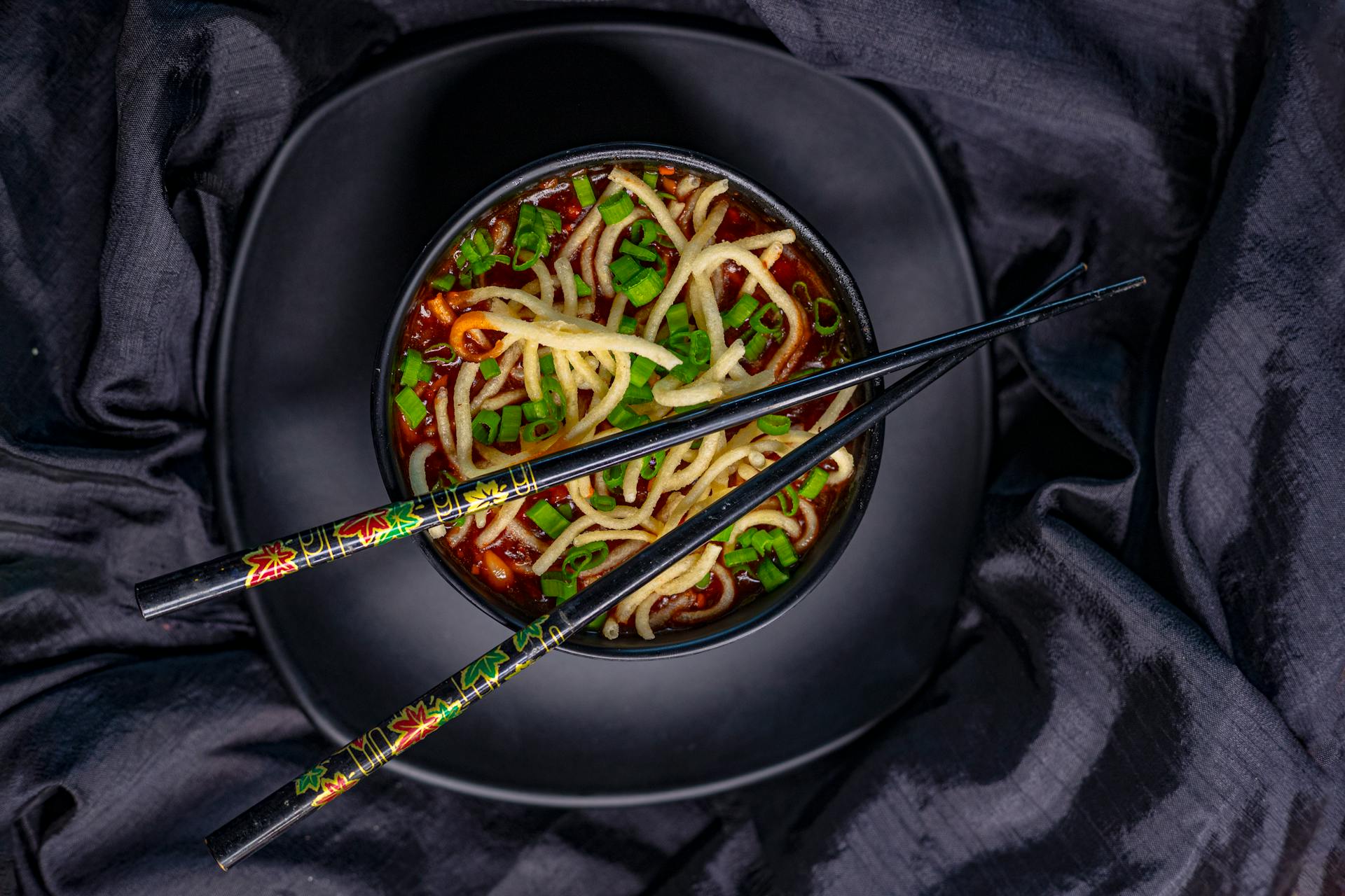 Top view of black bowl with Asian soup with noodle and green onion served with bamboo chopsticks on black tablecloth