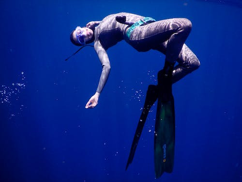 Side view of graceful young female diver in wetsuit flippers and mask swimming under clear deep blue ocean water in sunlight