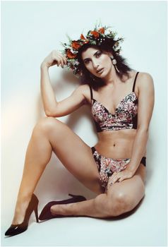 A stylish woman in floral lingerie and heels posing with a flower crown against a white background.