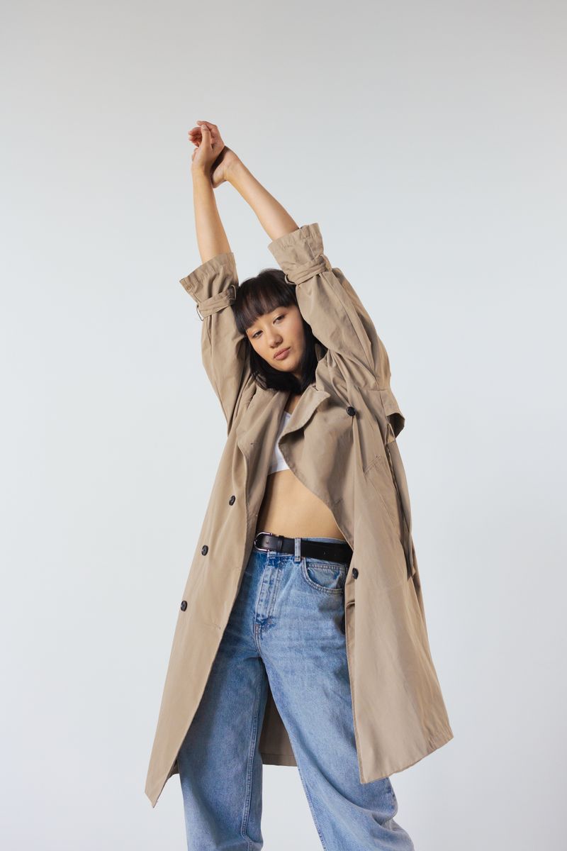 A person in a tan trench coat stretching both arms overhead against a pale studio backdrop — photo by cottonbro studio on Pexels