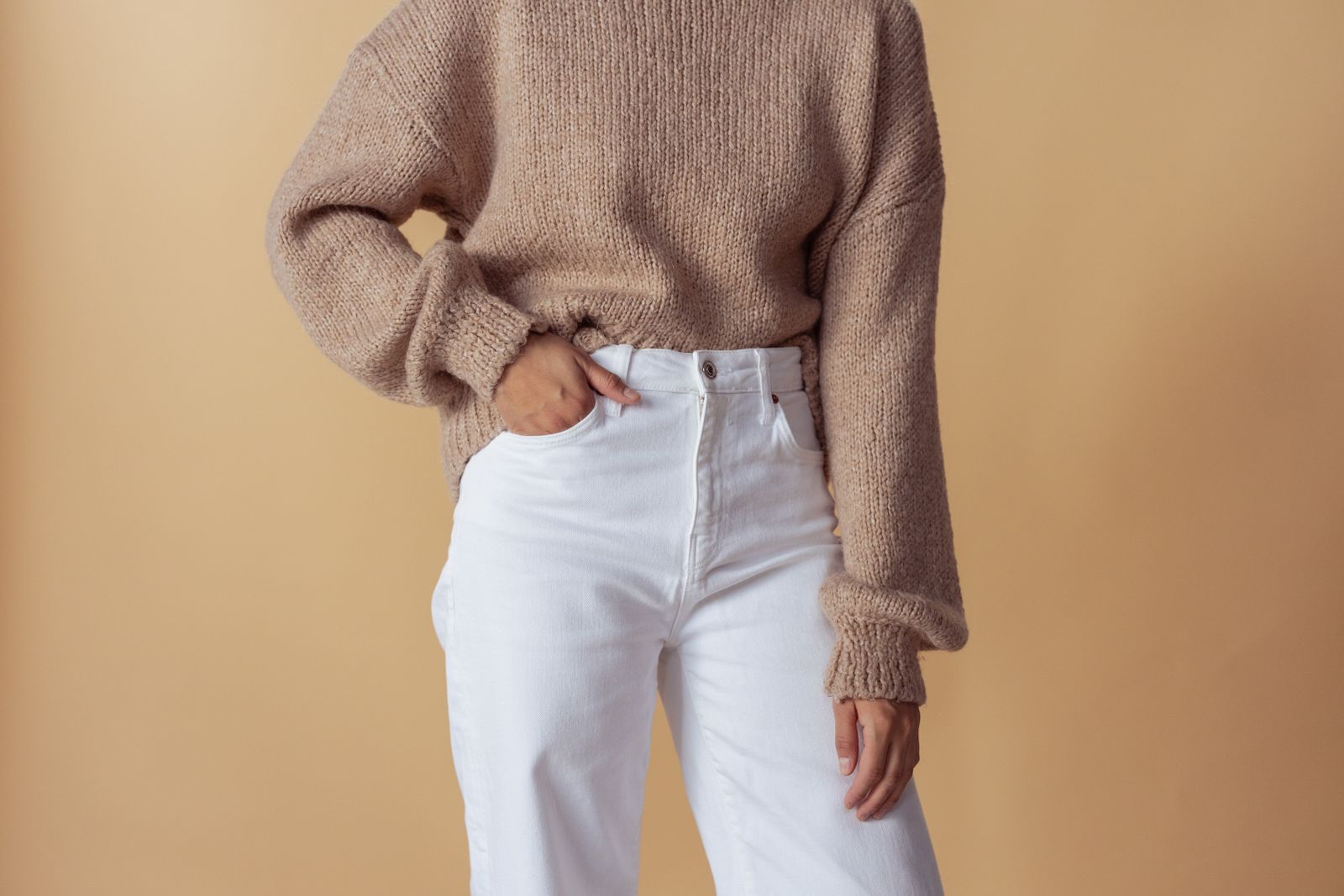 Woman in a tan knit sweater and white high-waisted jeans, hand on hip, against a beige backdrop