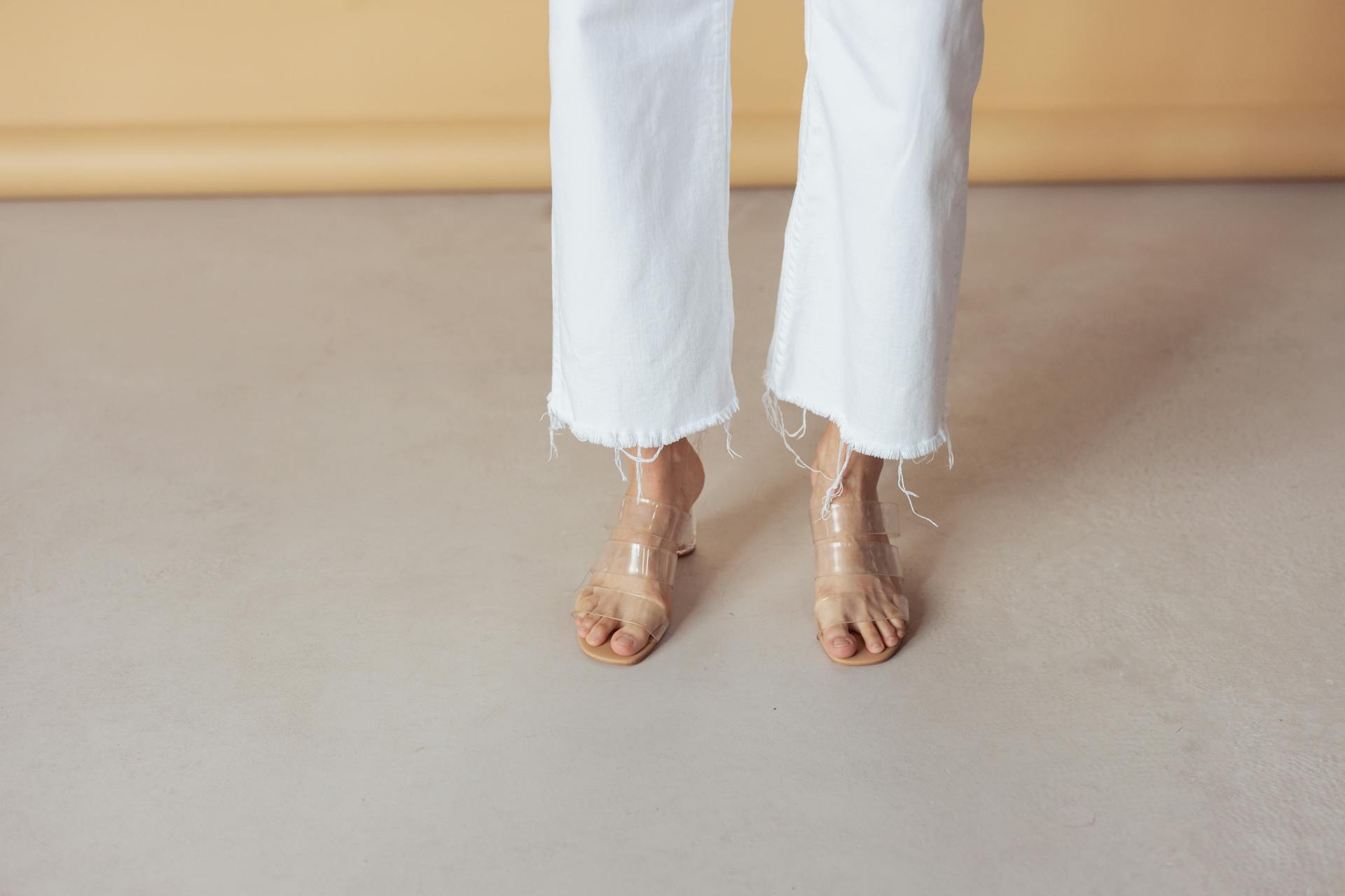 A close-up of feet in transparent sandals paired with white frayed pants on a neutral background.