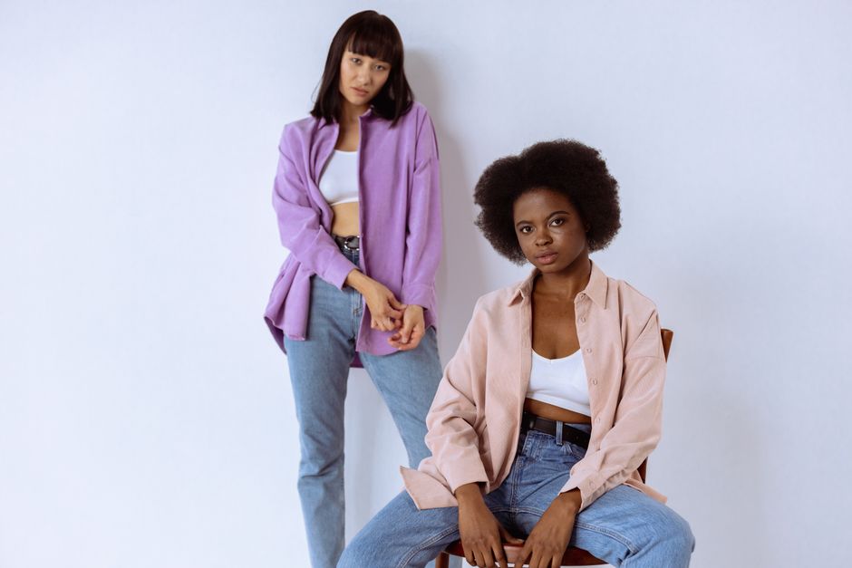 Two stylish women pose in a casual studio setting showcasing trendy fashion with pastel shirts.
