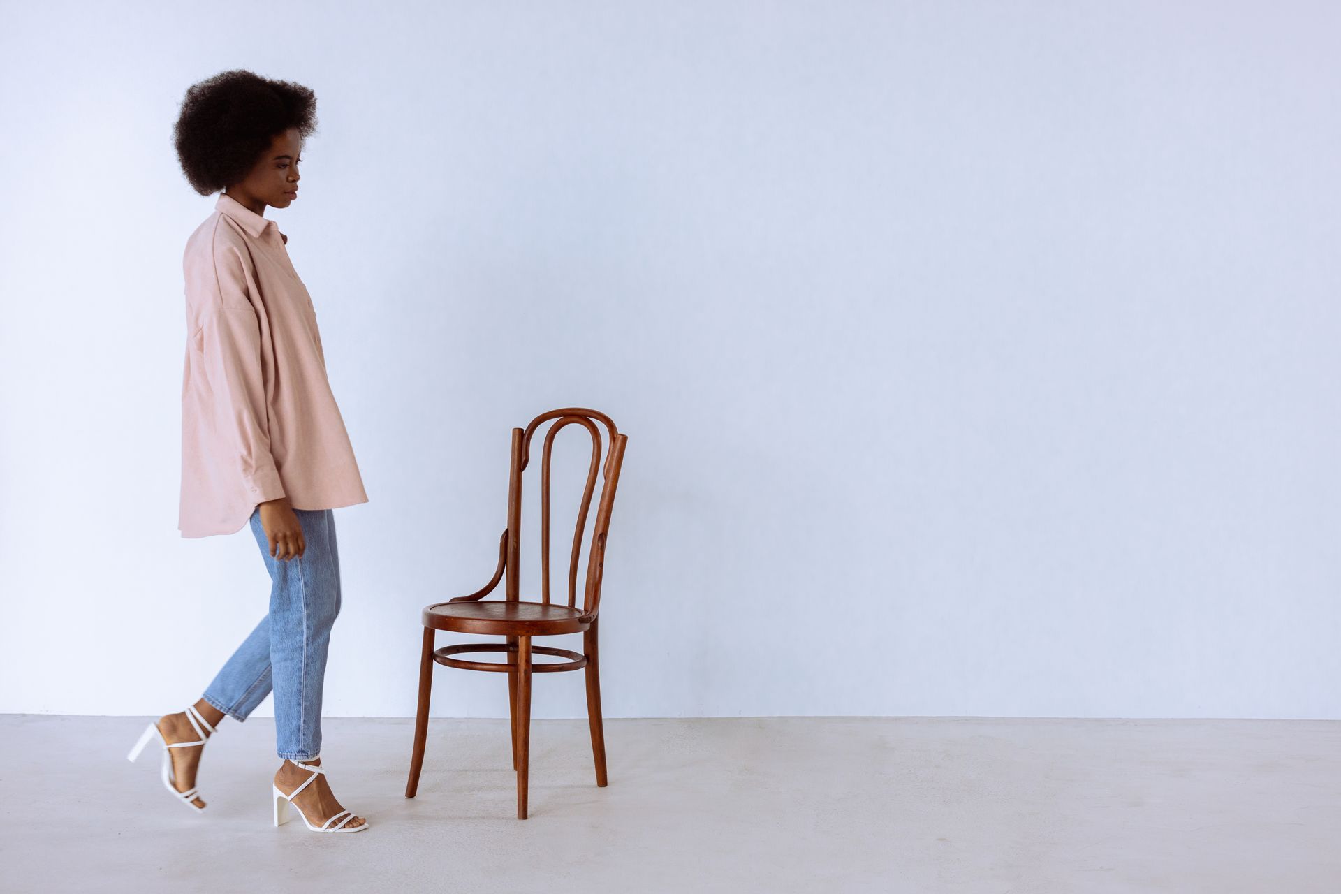 A Woman in Pink Dress Shirt and Blue Denim Jeans Walking Towards a Wooden Chair