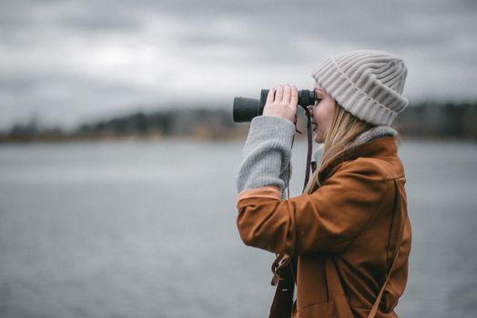 Side view of female in warm clothes smiling and using binoculars on blurred background of embankment