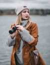 Young female with leather handbag and binoculars looking away on blurred background on gloomy river in cold weather on blurred background