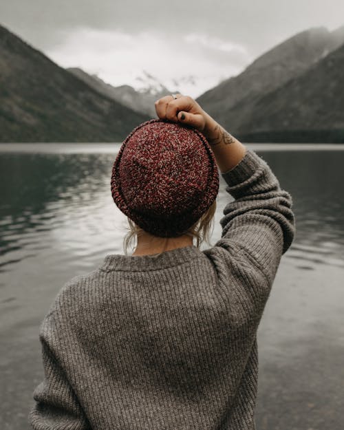 Tattooed hipster in woolen red hat touching head near mountains