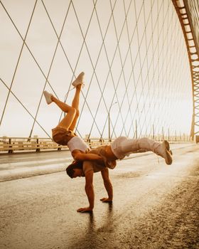 Full body of energetic anonymous man and woman doing counterbalance pose of dance on asphalt road of huge bridge in sunlight