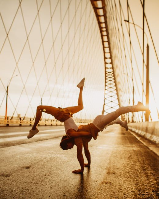 Full body of man doing push up jump while performing counterbalance pose with woman dancing on sunny wet asphalt road of massive bridge